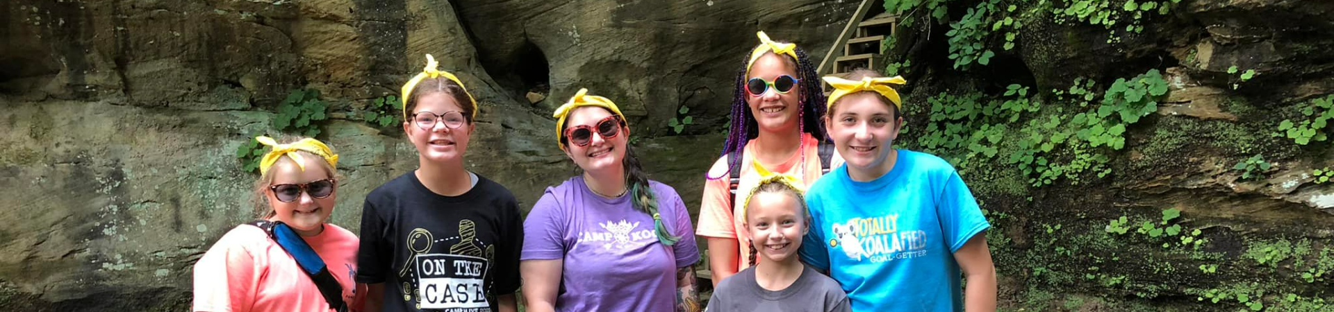  six girls all in yellow bandana headbands standing against a rock wall smiling 